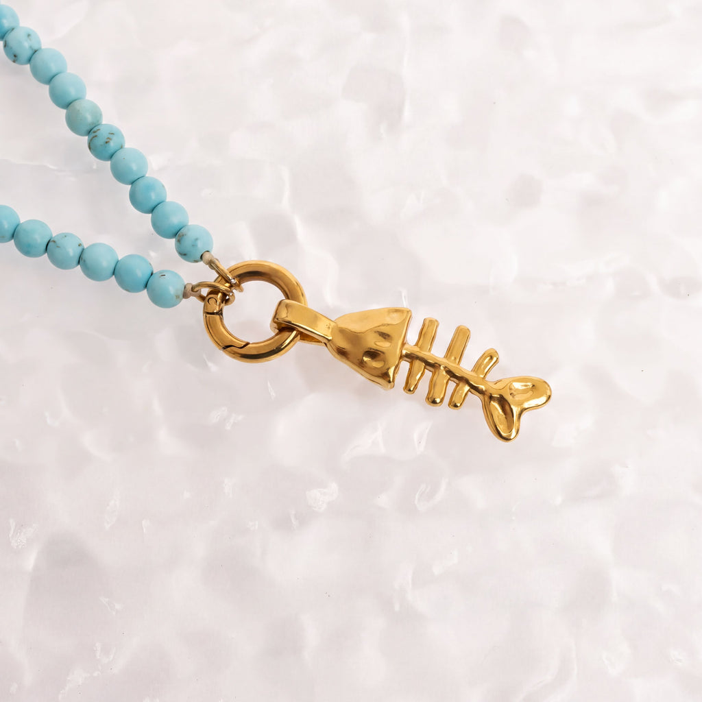 A gold-colored fish-shaped pendant with turquoise beads is displayed against a white textured background.