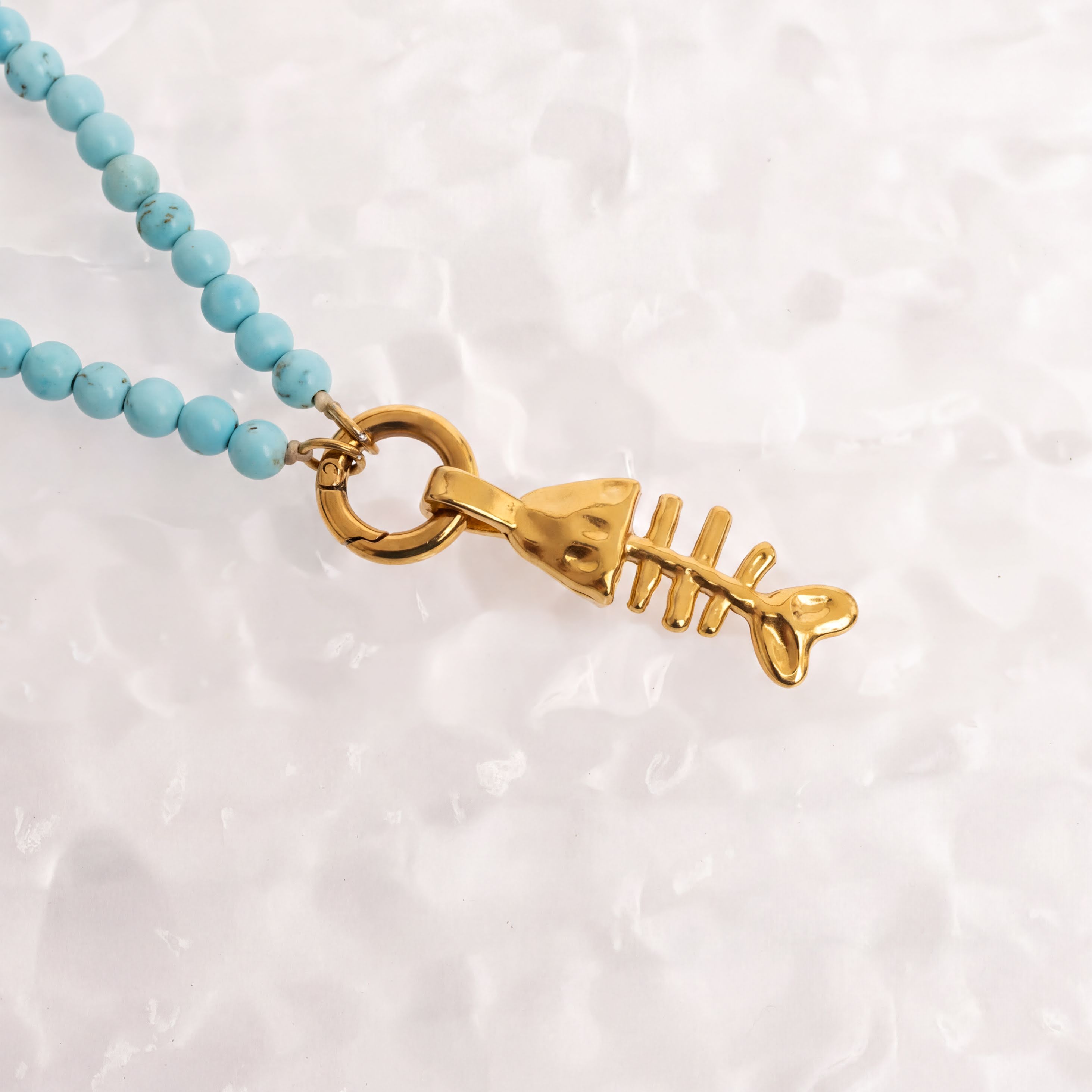 A gold-colored fish-shaped pendant with turquoise beads is displayed against a white textured background.