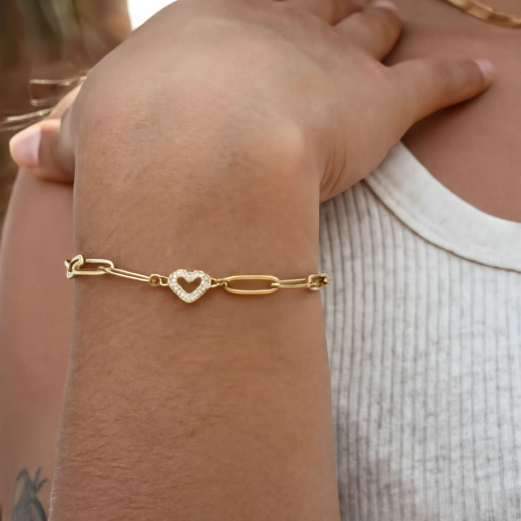 A close-up view of a person's wrist wearing a gold-colored bracelet with a heart-shaped charm.