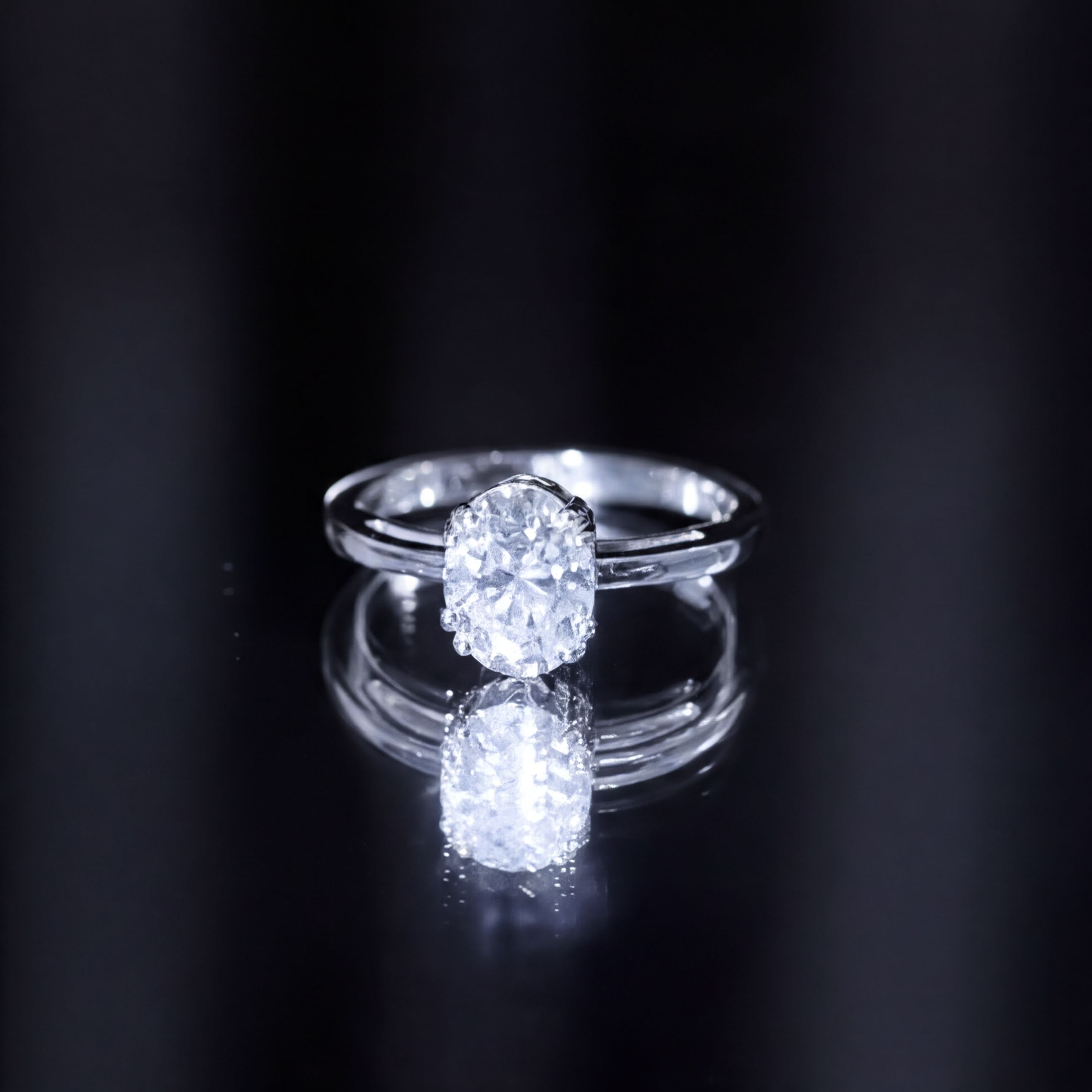 A close-up of a diamond engagement ring with a large central diamond set in a silver band, reflecting on a dark surface.