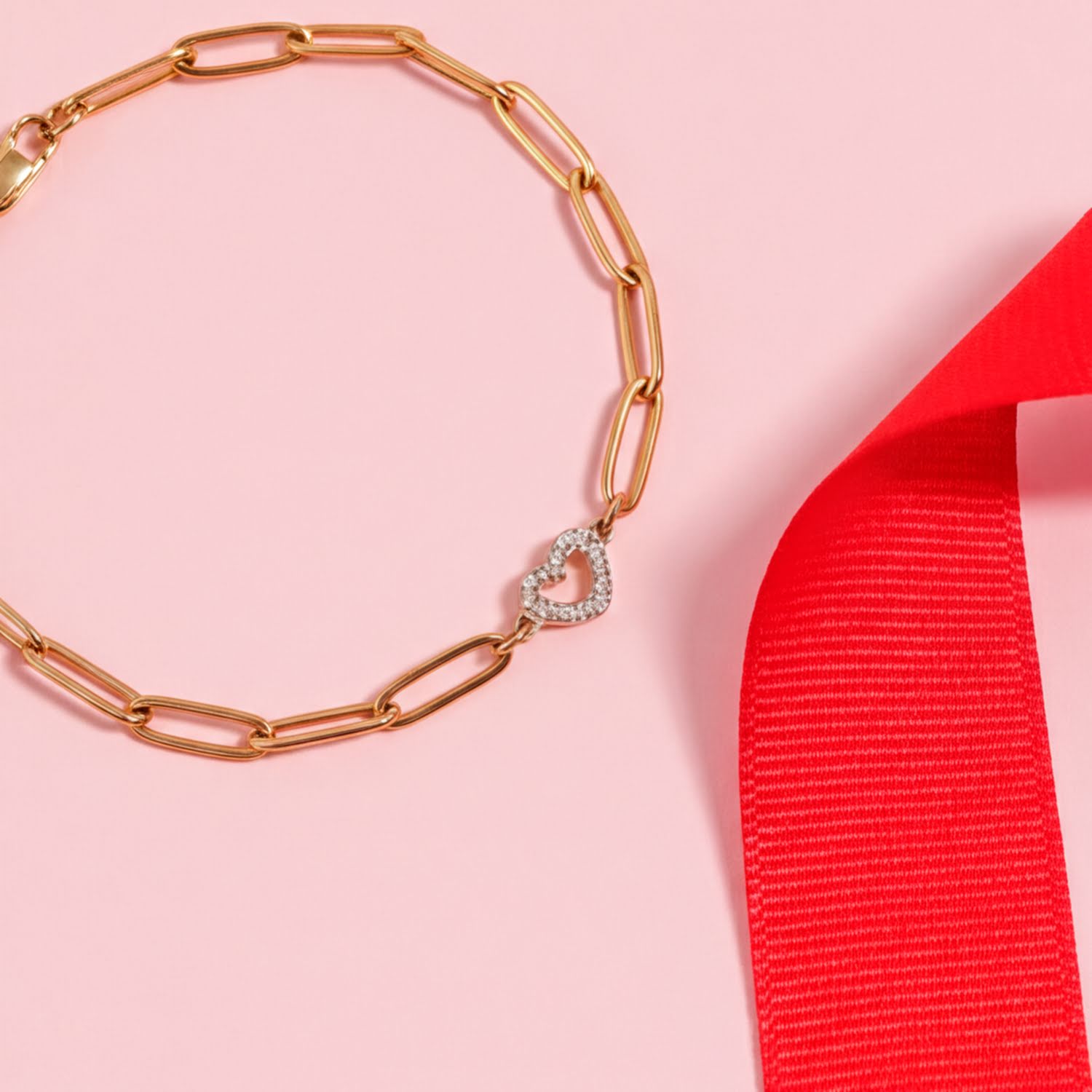 A gold-colored bracelet with a heart-shaped charm is displayed on a pink background, accompanied by a red ribbon.