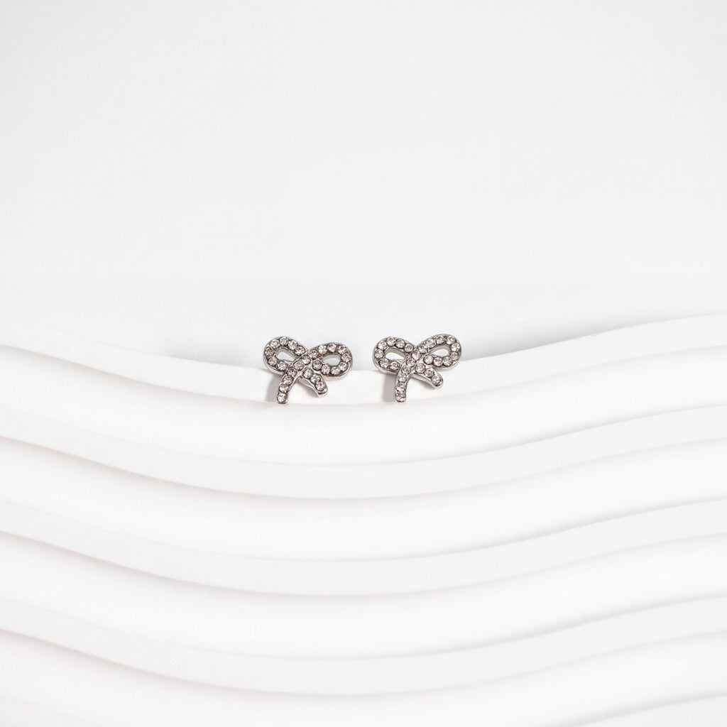 A pair of silver earrings with a bow design, placed on a white surface with a wavy pattern.