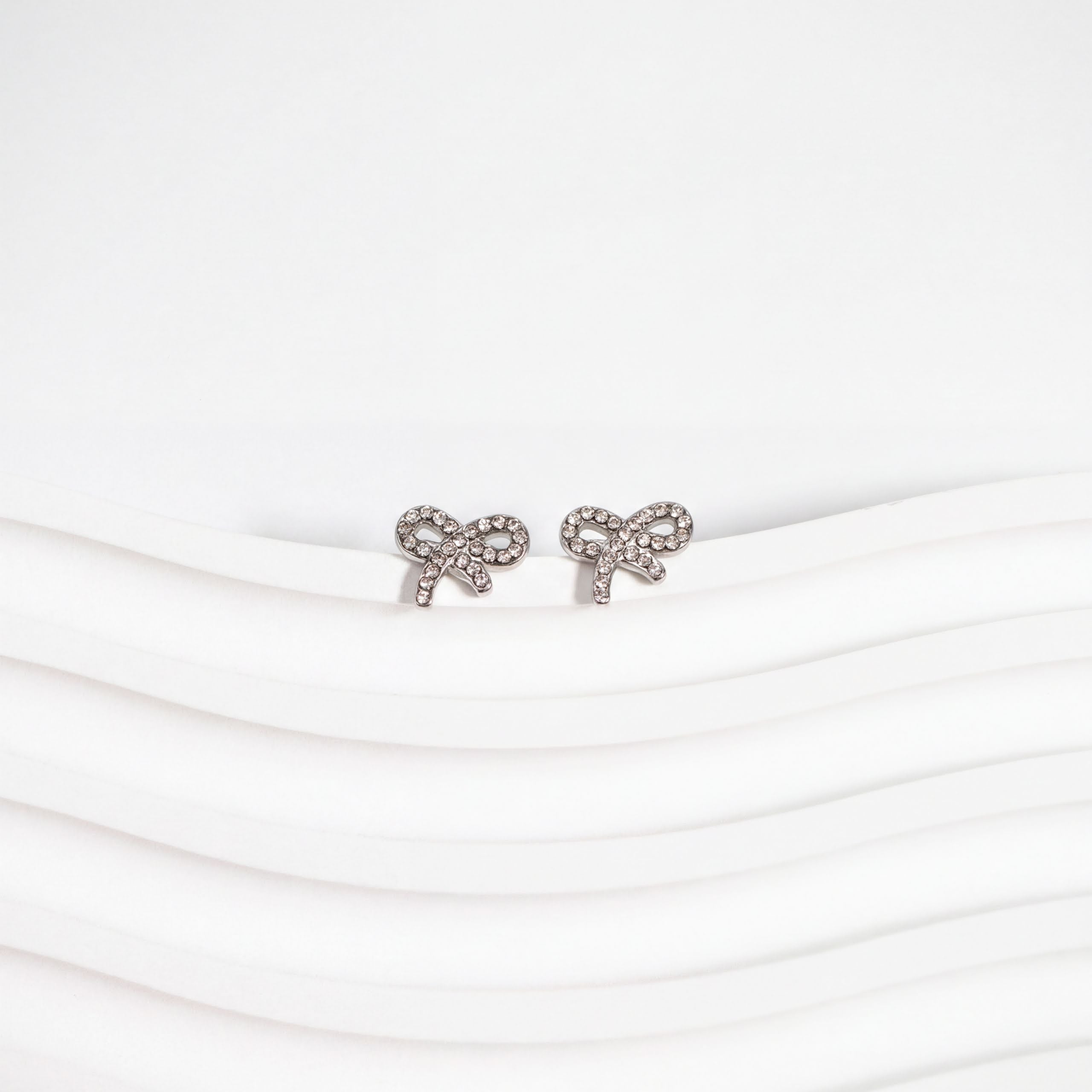 A pair of silver earrings with a bow design, placed on a white surface with a wavy pattern.