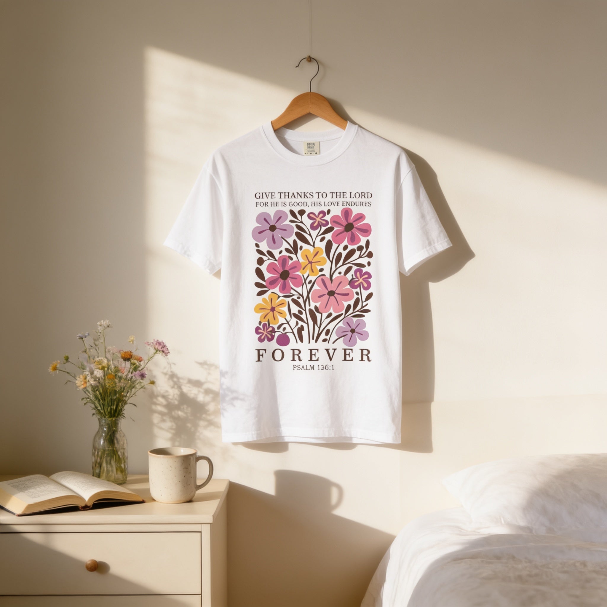 A white t-shirt with a floral design hangs on a wooden hanger, accompanied by a vase of flowers, a mug, and a book on a dresser.
