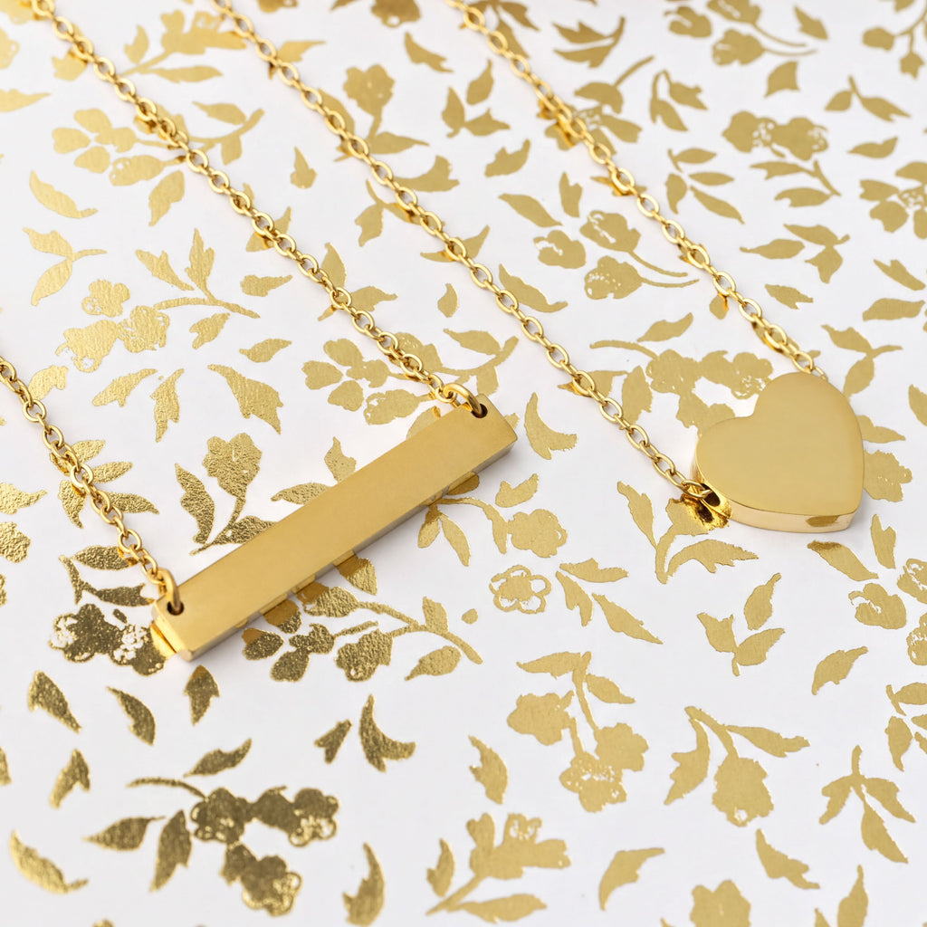 Two gold necklaces, one bar-shaped and one heart-shaped, on a gold floral patterned background.
