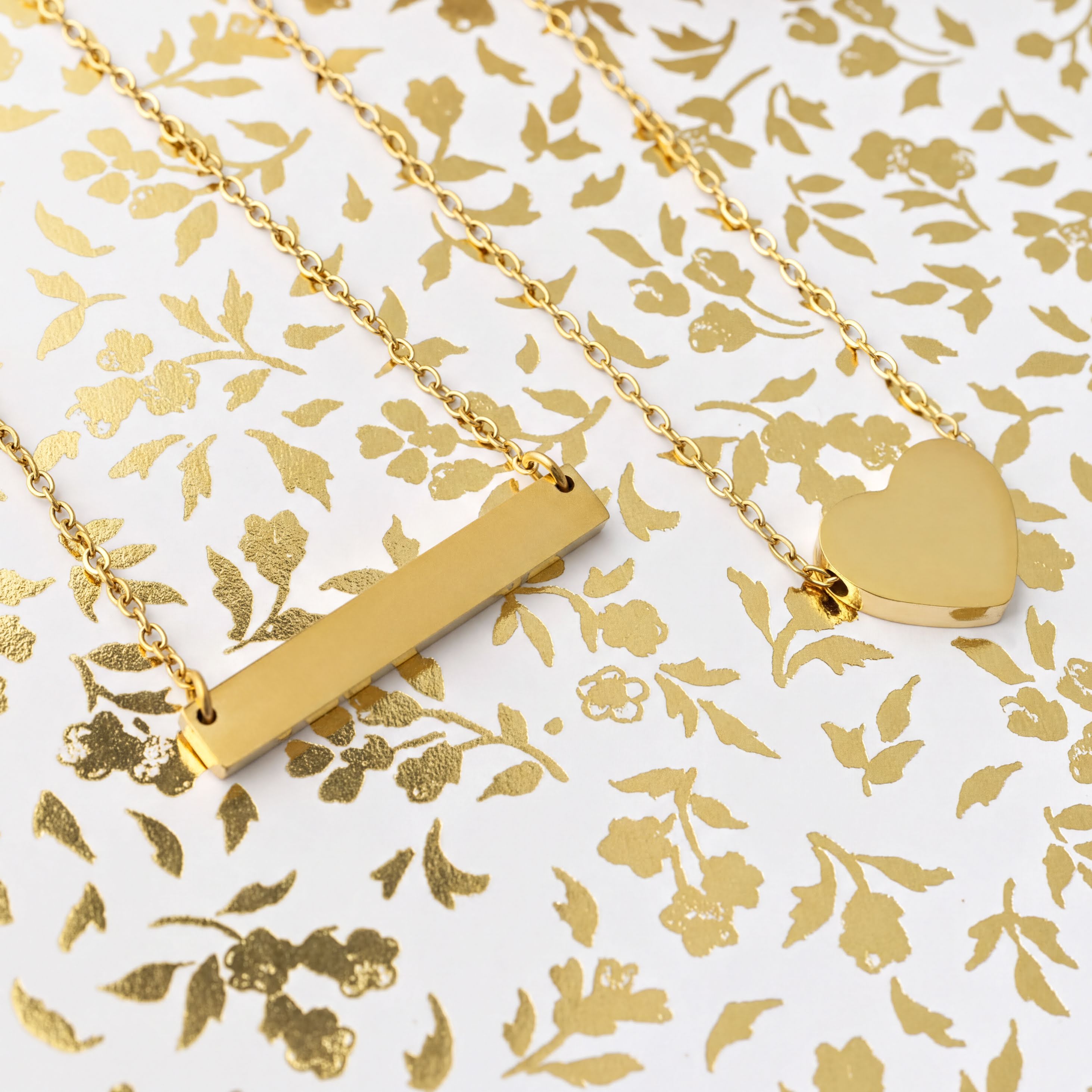 Two gold necklaces, one bar-shaped and one heart-shaped, on a gold floral patterned background.