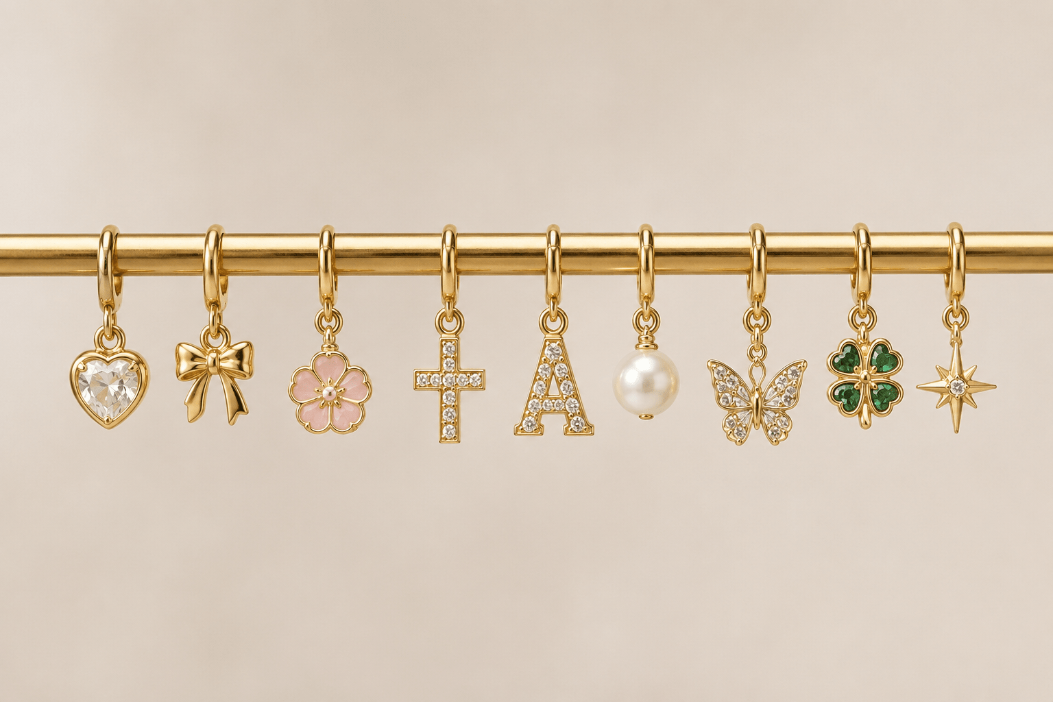 A row of gold-tone charm hooks or pendants hanging from a gold rod against a warm beige background. The charms include a heart with a clear stone, a bow, a pink flower, a cross, an outlined letter A, a pearl bead, a butterfly with stones, a green four-leaf clover, and a star.
