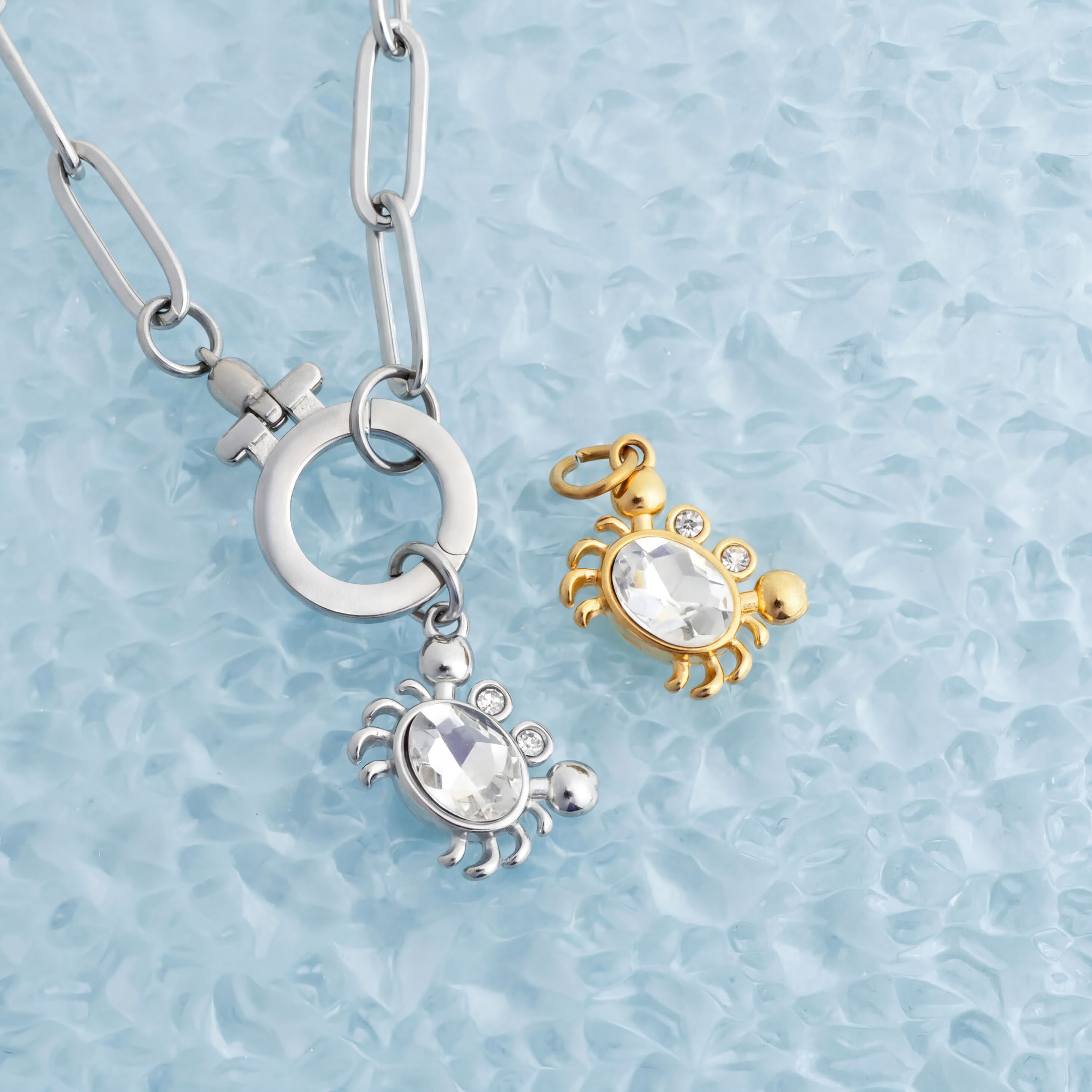 two pieces of jewelry, one silver and one gold, with clear gemstones attached to them. The silver piece has a round charm with a lobster design, while the gold piece has a lobster design as well.