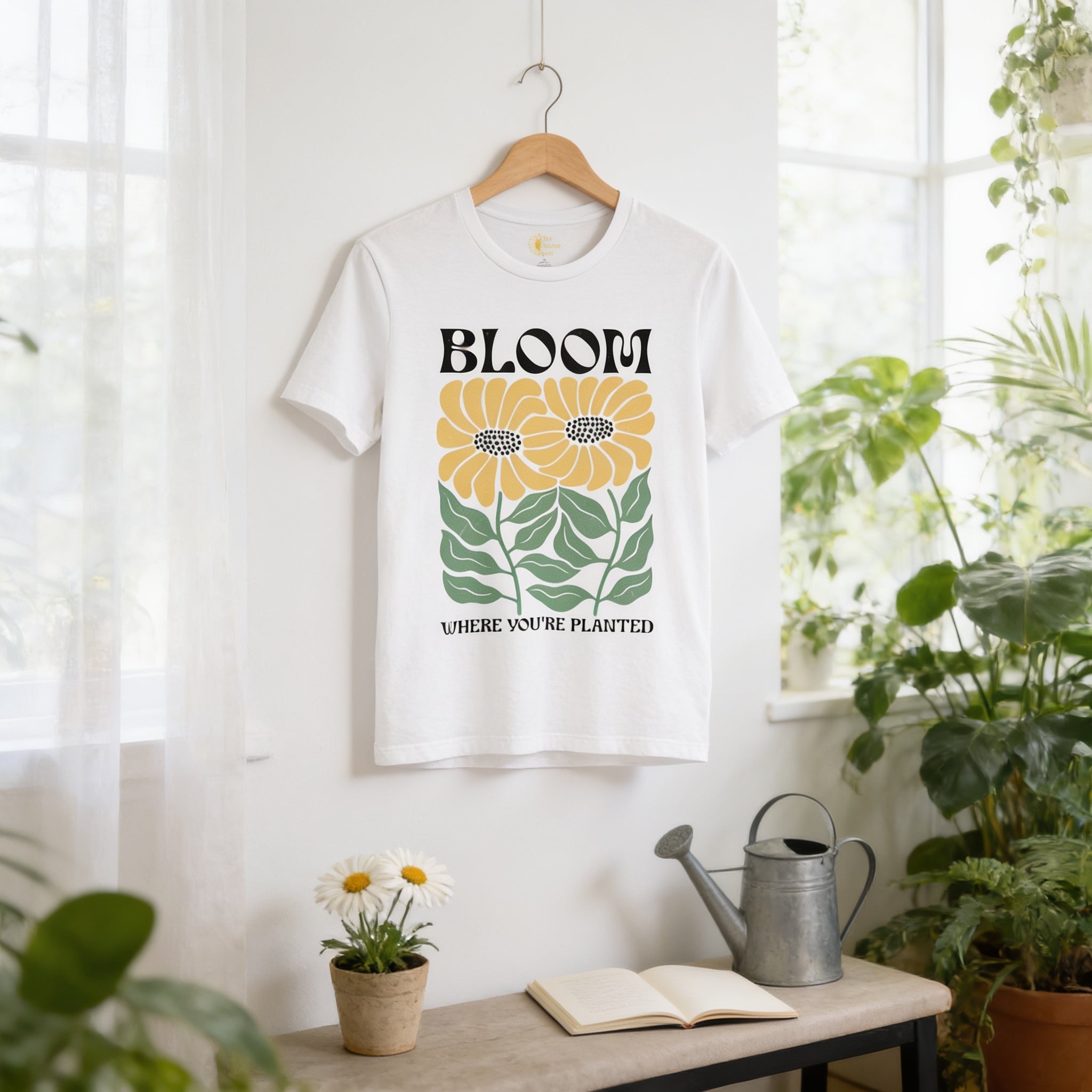 a white t-shirt with a graphic design of yellow flowers and green leaves, hanging on a wooden hanger on a wall. The t-shirt has the text "BLOOM" and "WHERE YOU'RE PLANTED" printed on it. In the foreground, there is a potted plant, a watering can, and an open book on a table.