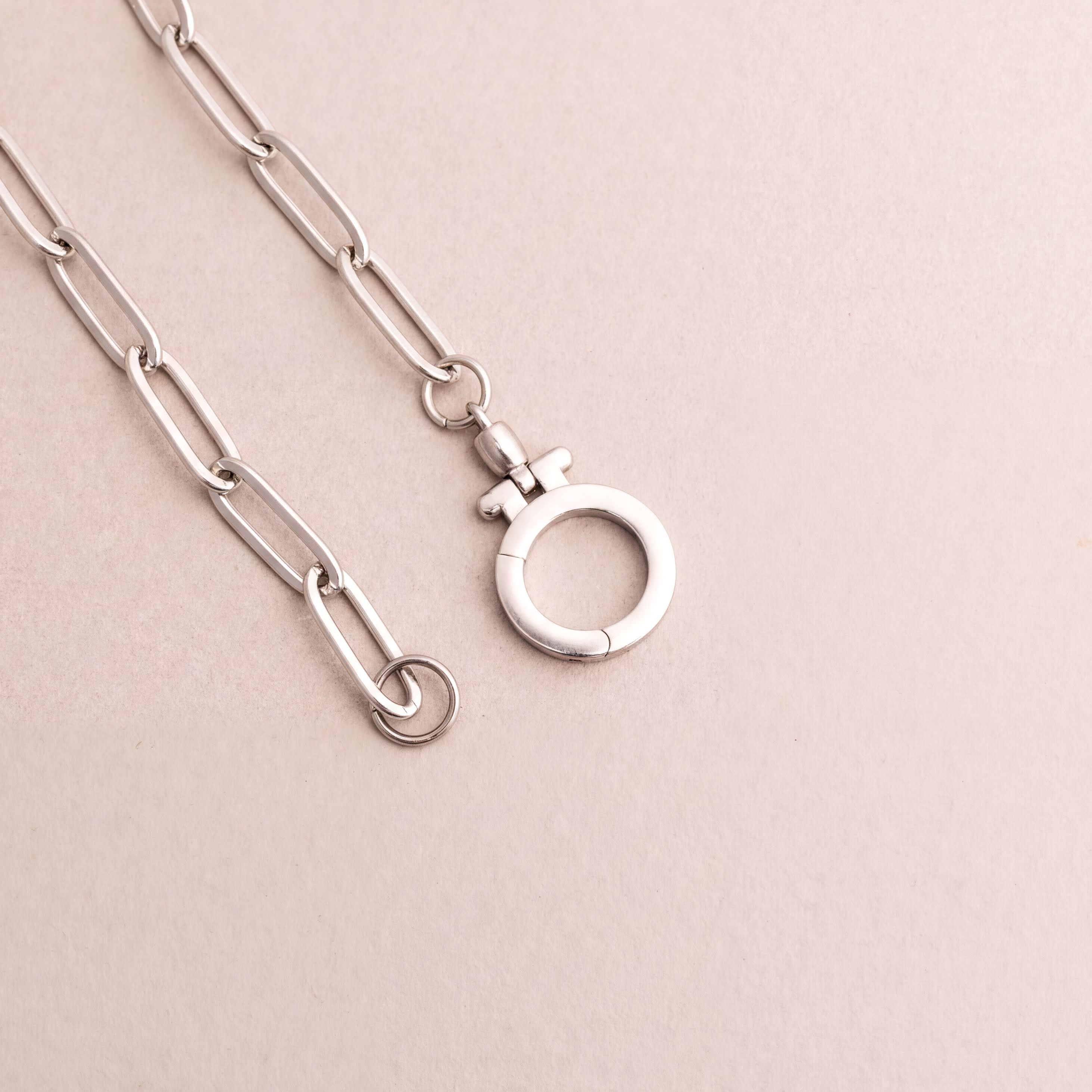 a silver chain with a round clasp attached to it, resting on a light pink background.