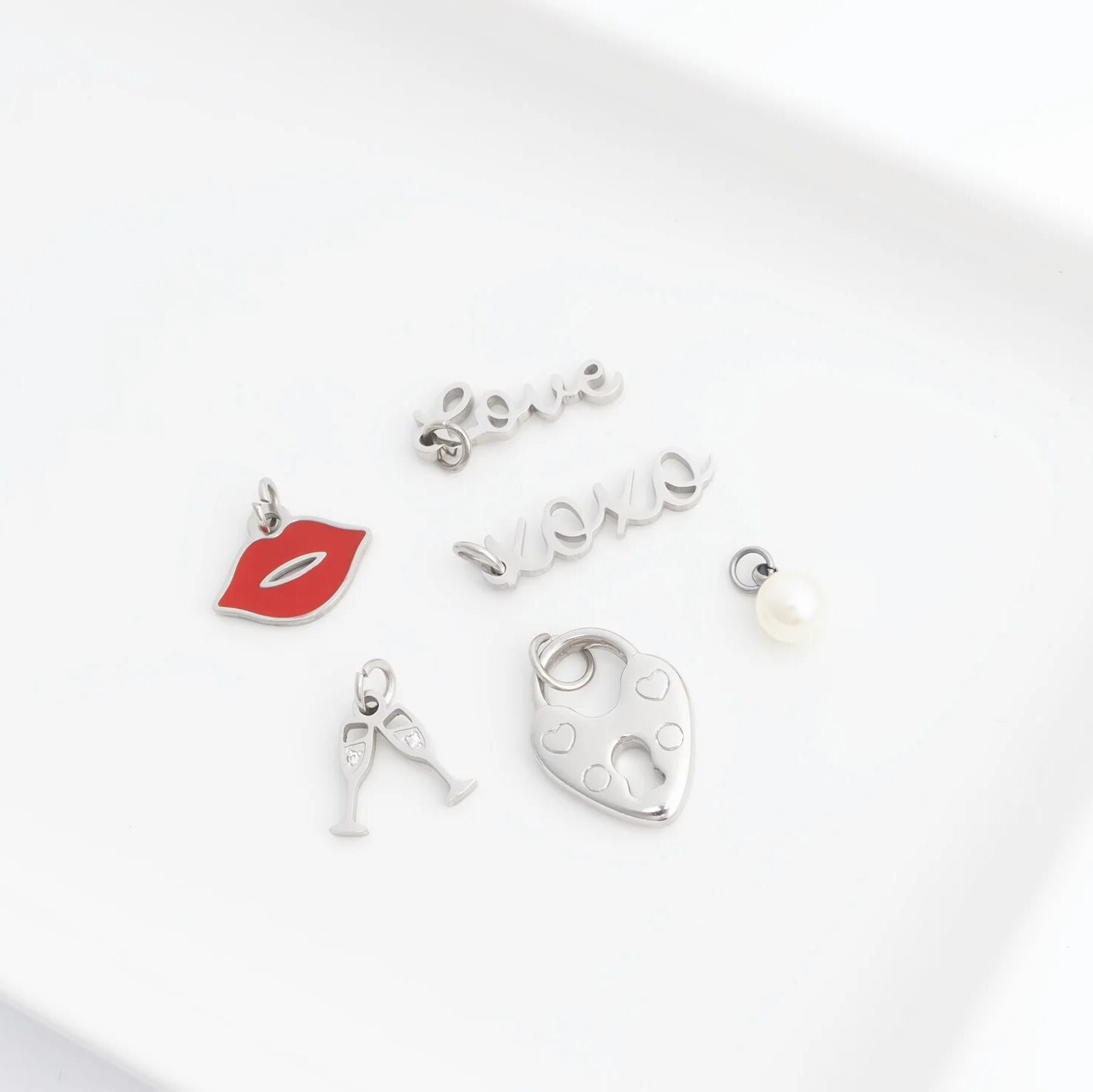 Collection of silver charms including a red lip, letter 'A', heart, and pearl on a white background