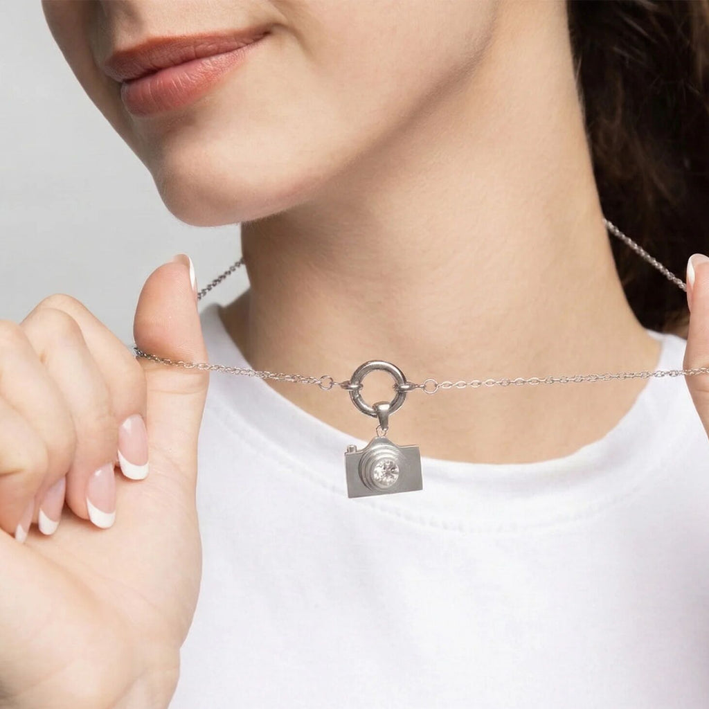 Person wearing a silver necklace with a camera-shaped pendant on a plain background
