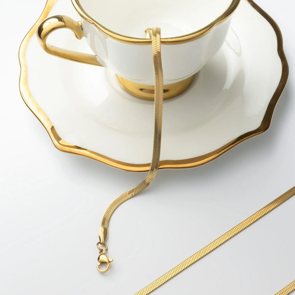Gold necklace draped over a white teacup and saucer on a light gray background