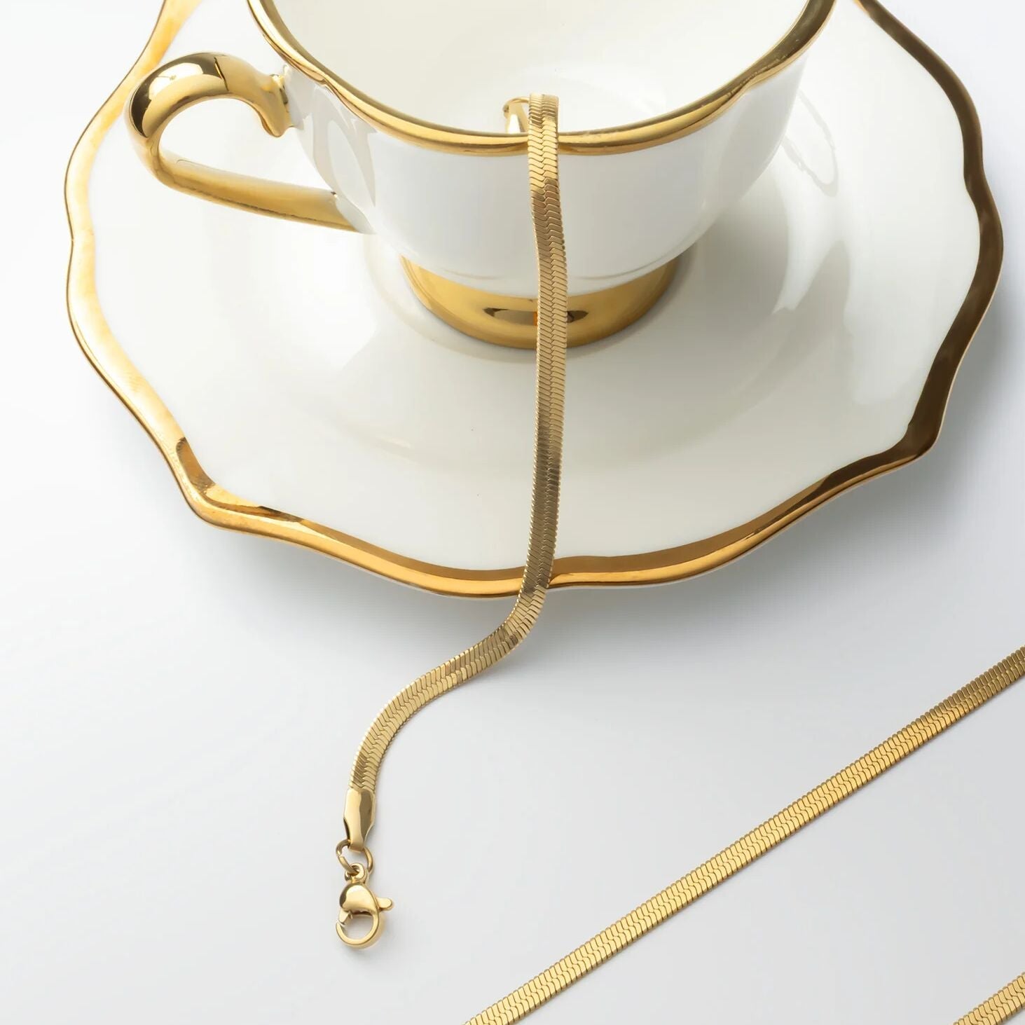 Gold necklace draped over a white teacup and saucer on a light gray background