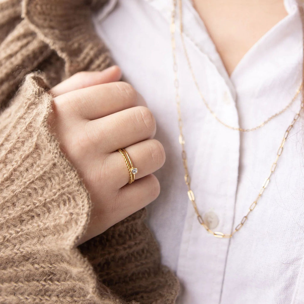 Close-up of a hand wearing a gold ring with a diamond, holding a brown textured sleeve, with a white shirt in the background.