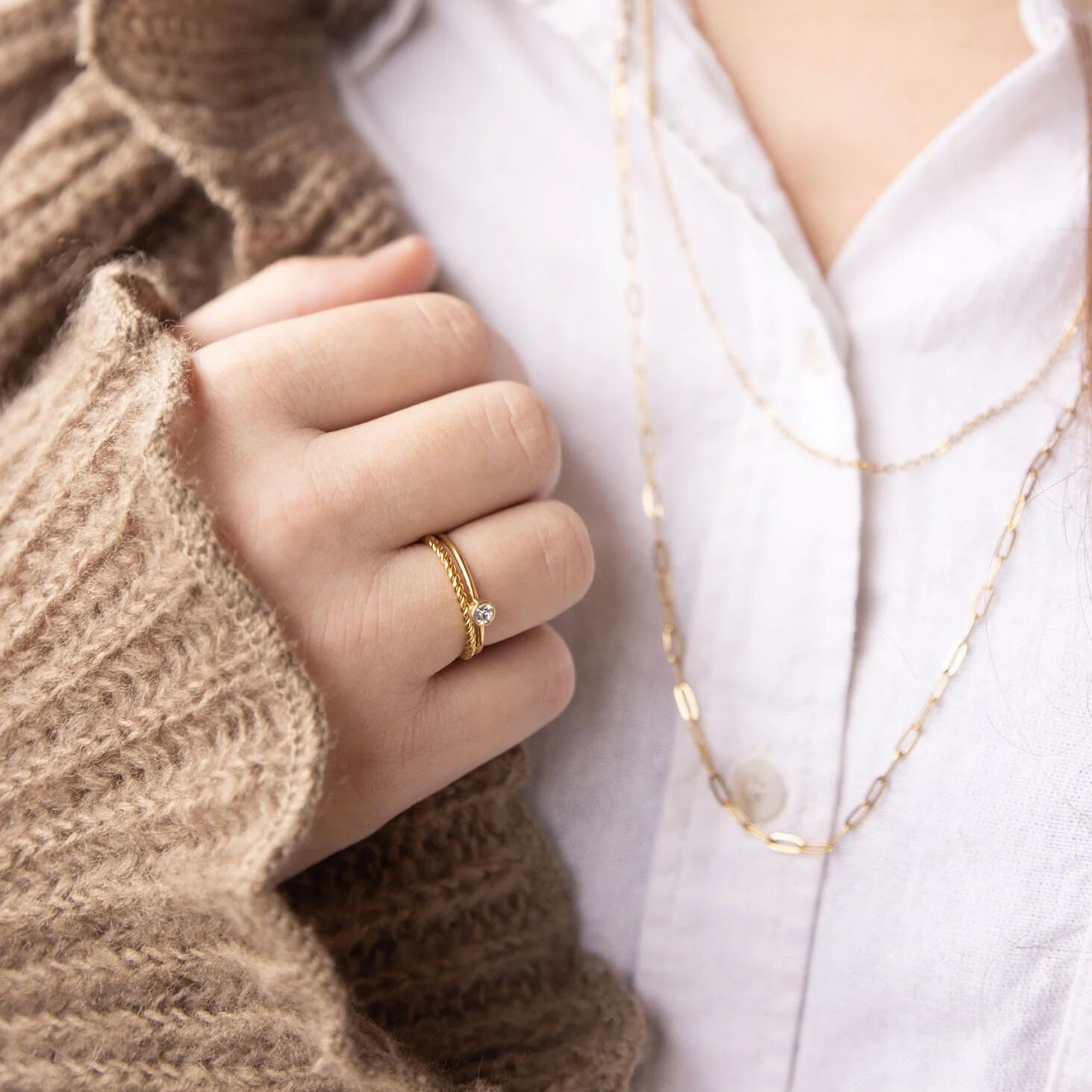 Close-up of a hand wearing a gold ring with a diamond, holding a brown textured sleeve, with a white shirt in the background.