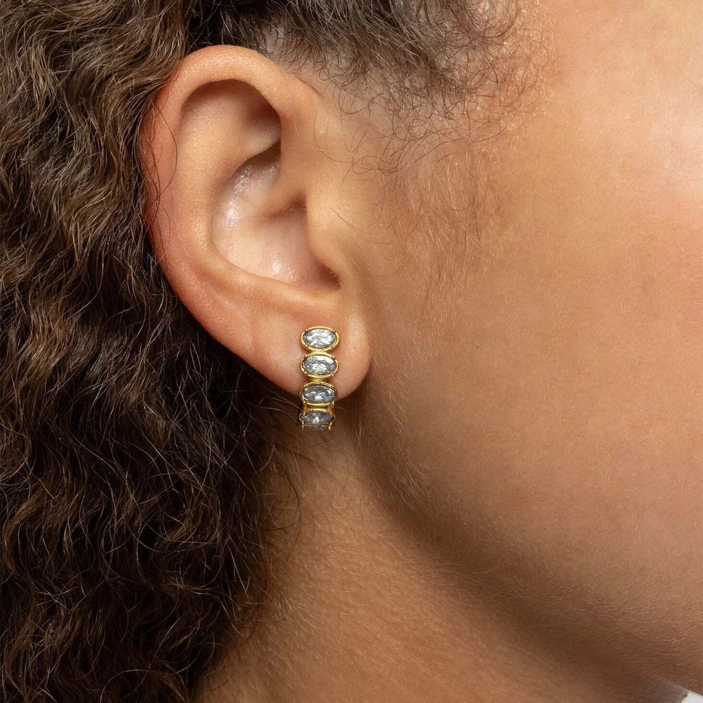 Close-up of an ear wearing a gold Oval Stone Huggie Earrings with clear stones.