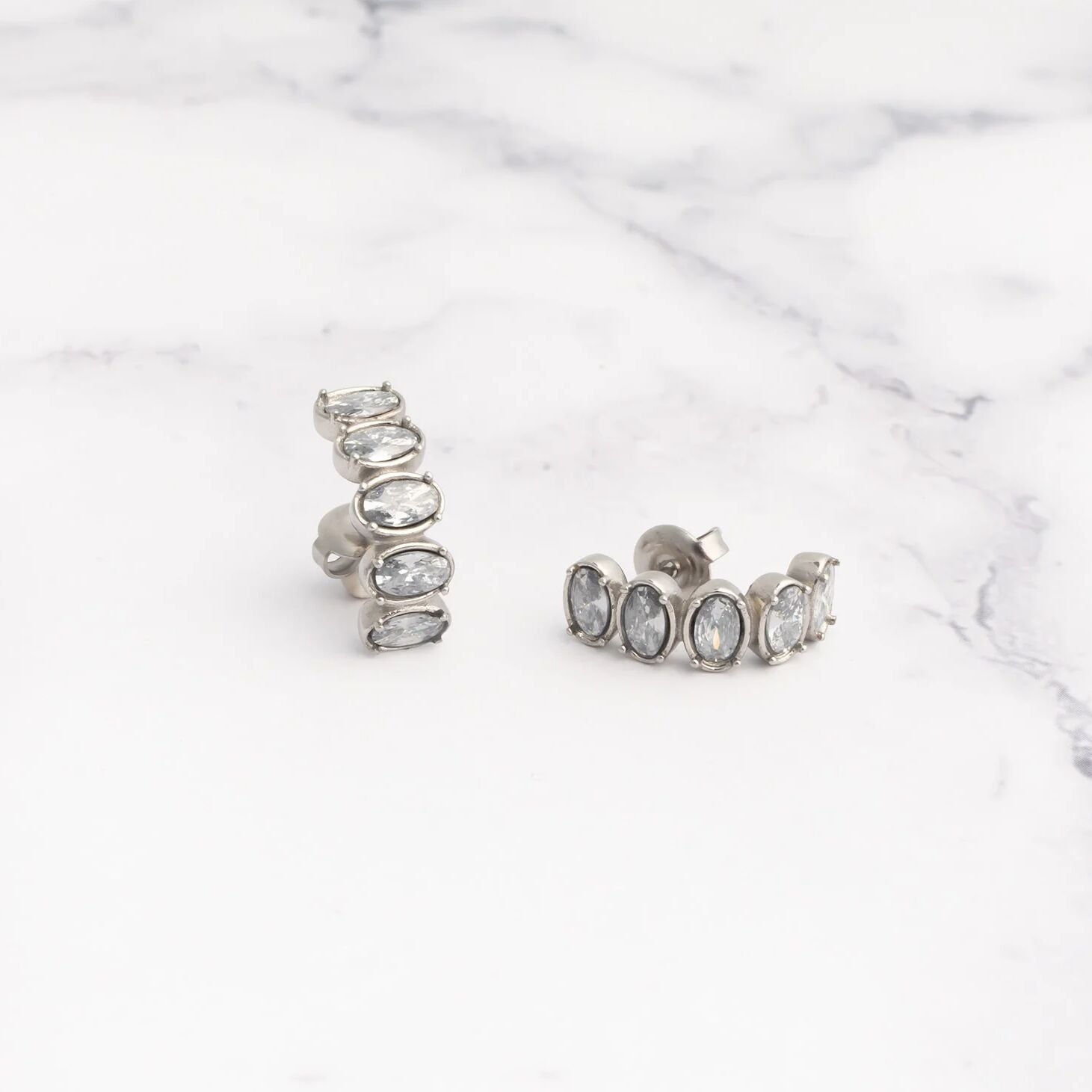 Pair of silver hoop Oval Stone Huggie Earrings with clear gemstones on a marble surface