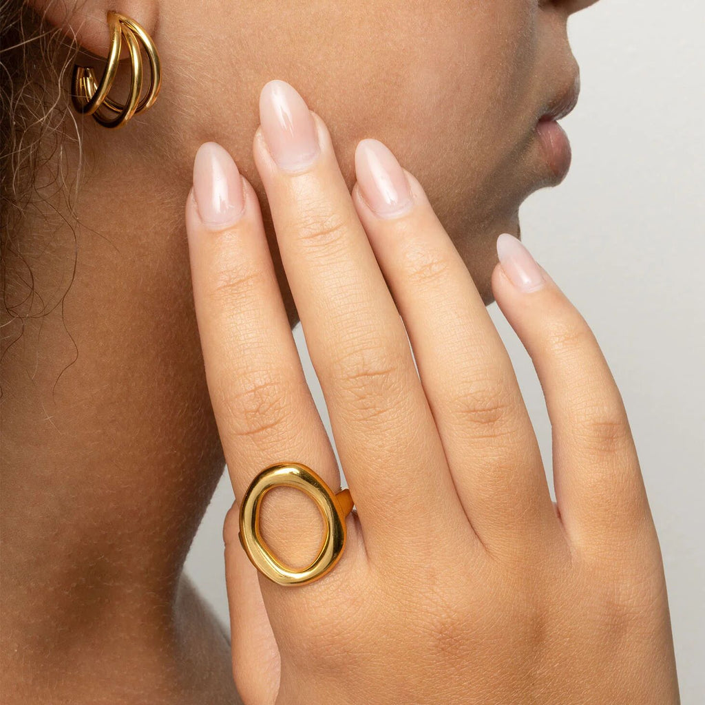 Close-up of a person wearing gold hoop earrings and a gold Oval Motif Ring  on a neutral background