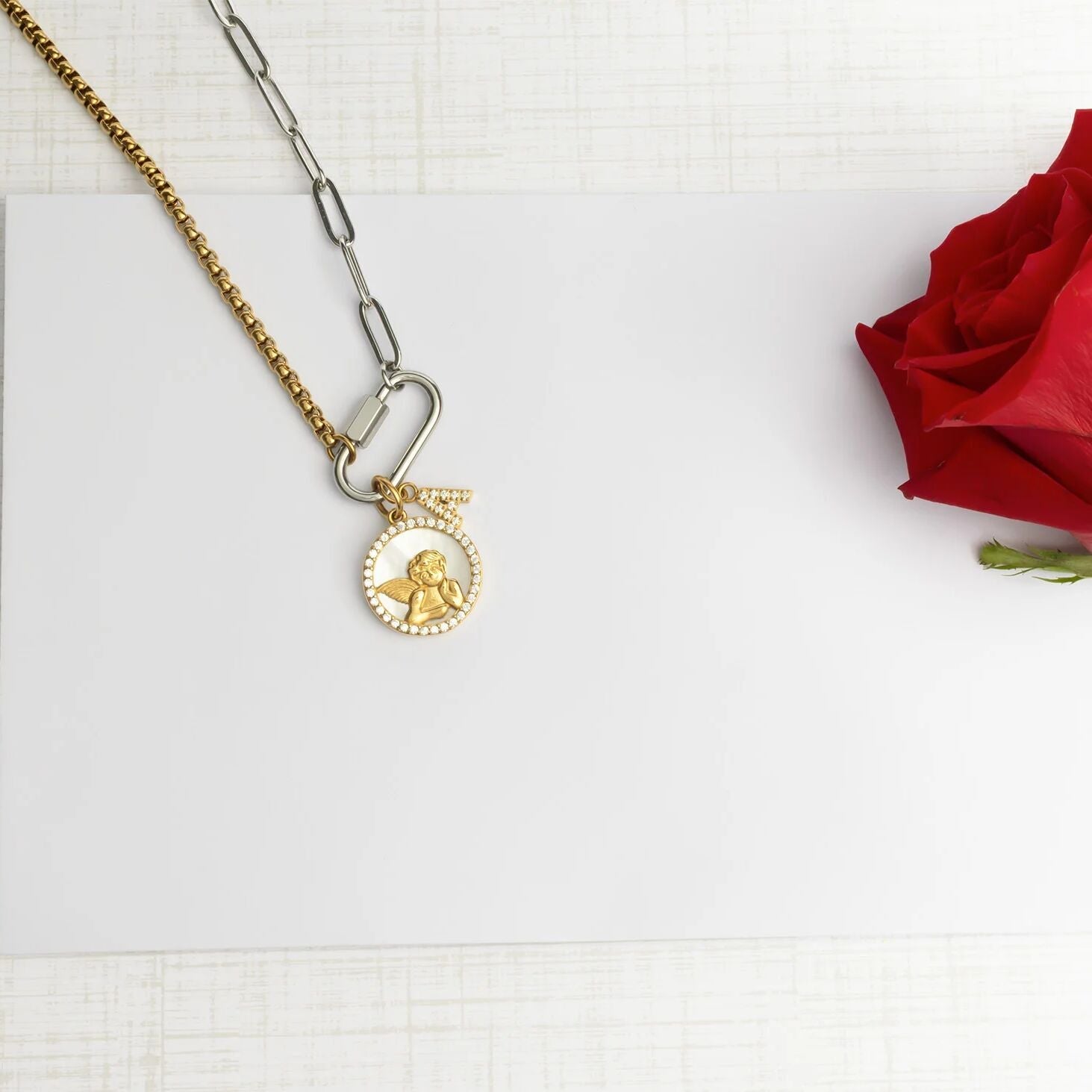 Gold necklace with a pendant on a white surface next to a red rose.