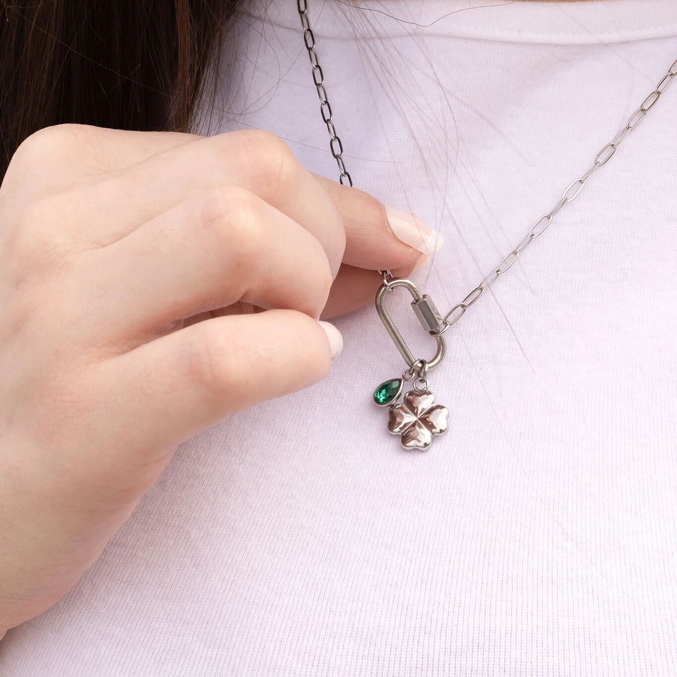 Hand holding a silver necklace with a clover charm and green gemstone against a white background