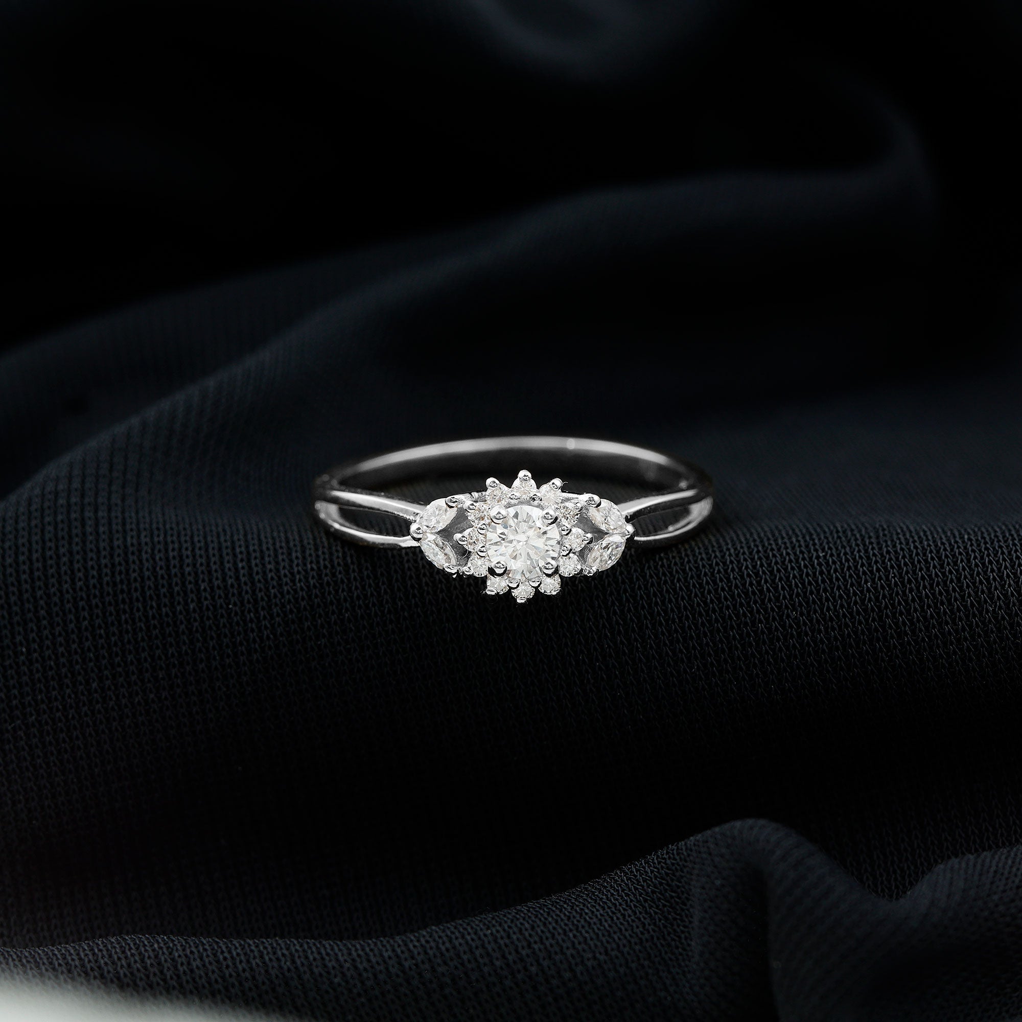 A diamond engagement ring with a large central diamond surrounded by smaller diamonds is displayed on a dark fabric background.