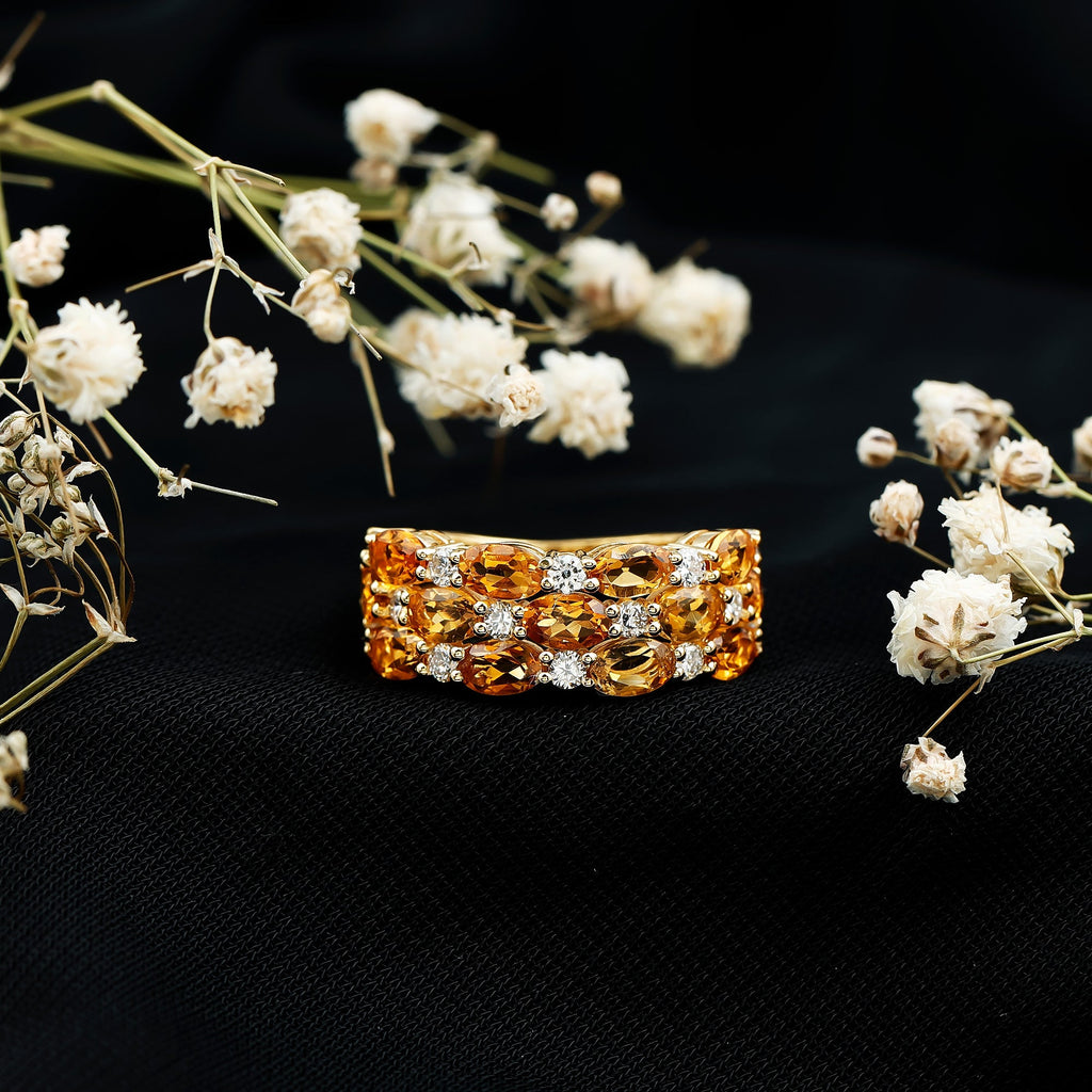 A gold ring with a large, ornate gemstone set in a ring band is displayed against a black background, surrounded by delicate white flowers.