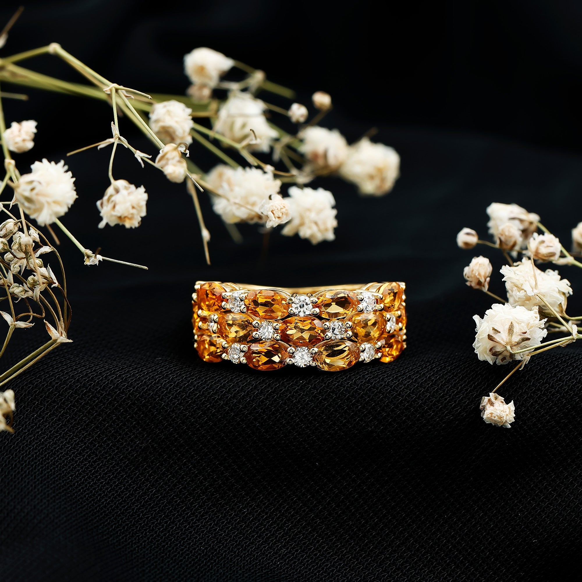 A gold ring with a large, ornate gemstone set in a ring band is displayed against a black background, surrounded by delicate white flowers.