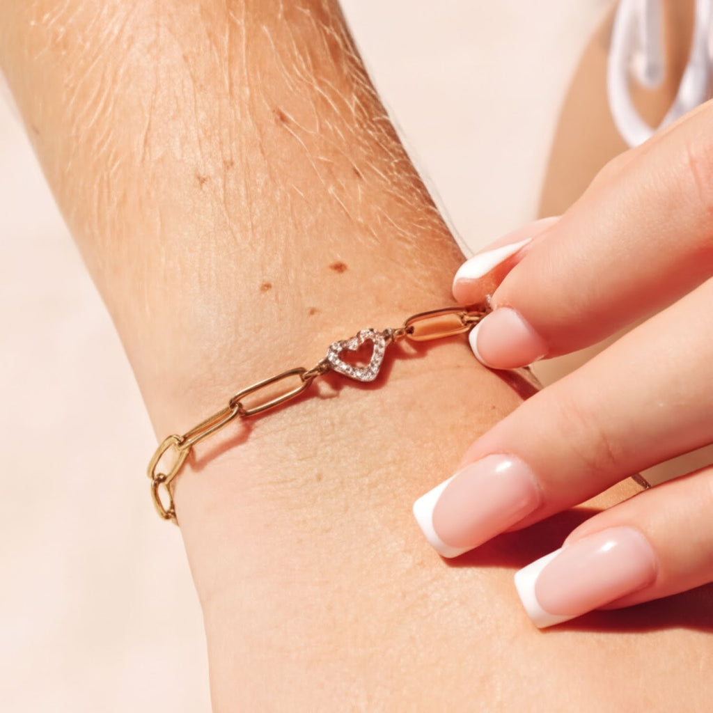A close-up view of a person's wrist wearing a gold-colored bracelet with a heart-shaped charm.