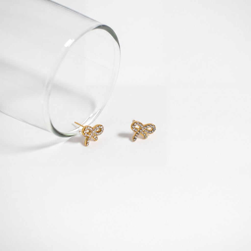 Two gold earrings with a bow design are placed on a white surface next to a clear glass.