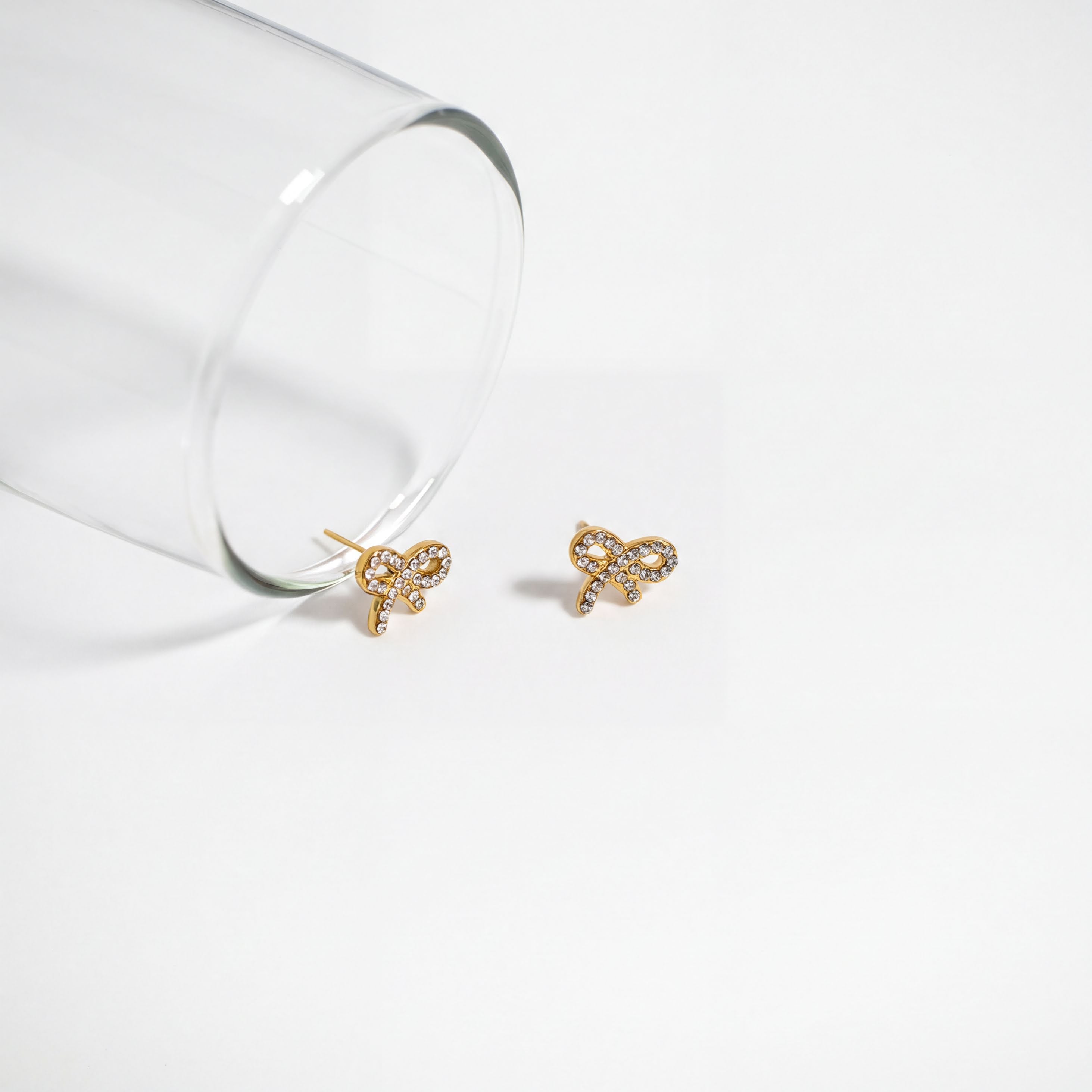 Two gold earrings with a bow design are placed on a white surface next to a clear glass.