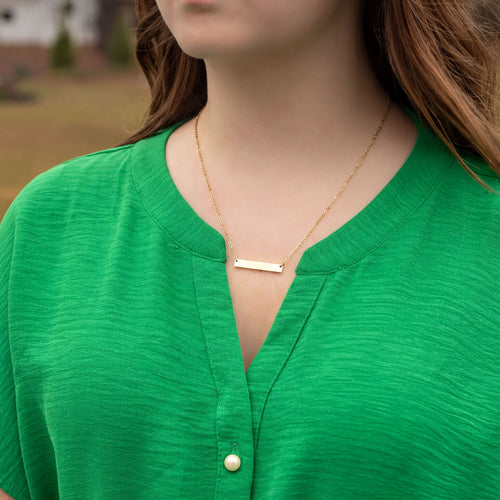 Person wearing a gold necklace with a bar pendant against a blurred outdoor background
