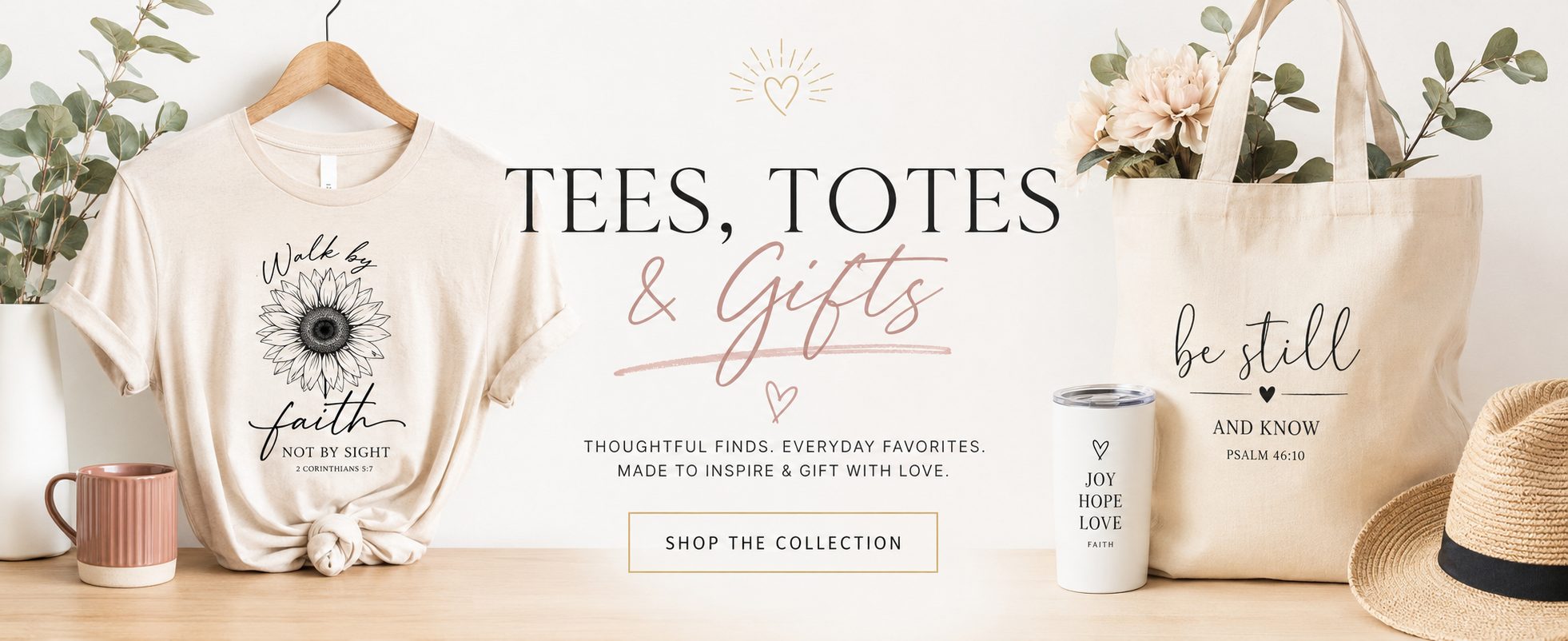 A styled collection banner featuring a cream graphic t-shirt on a wooden hanger, a beige tote bag with 'be still and know' text, a white can, and a straw hat on a light wood surface against a white wall.